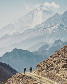 two people are walking on a trail in front of some mountains with snow capped peaks