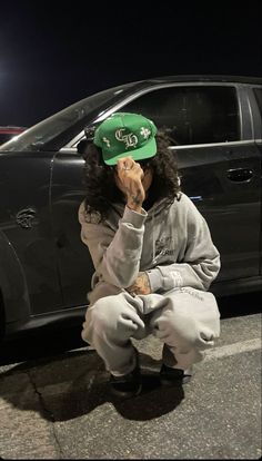 a person sitting on the ground in front of a car with a green hat over their face