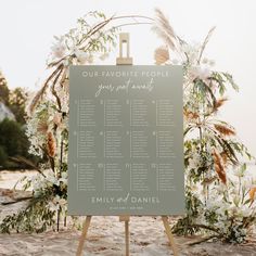 a sign that is sitting in the sand with flowers on it and greenery around it