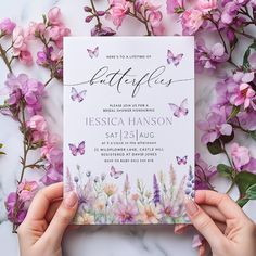 a person holding up a wedding card next to some pink flowers and purple butterflies on a marble surface