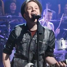 a man singing into a microphone while holding a guitar in front of him and other people behind him