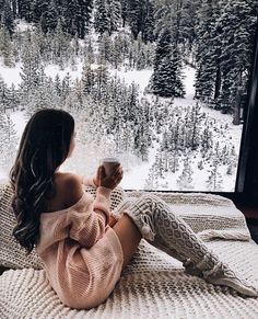 a woman sitting on top of a bed next to a window holding a coffee cup