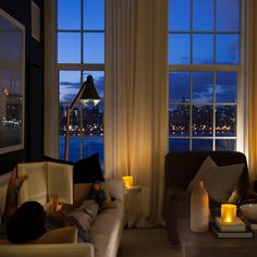 a person sitting on a couch reading a book in front of a window with the city lights lit up