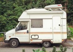 an rv parked on the side of the road near some trees and bushes with a surfboard mounted to it's roof