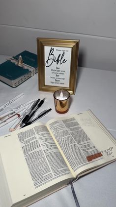 an open book sitting on top of a table next to a candle and some pens