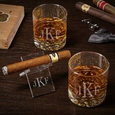 two glasses filled with whiskey and cigars on top of a wooden table