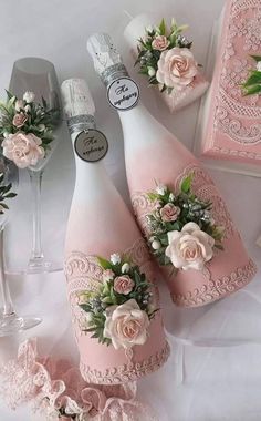 three pink wine bottles decorated with flowers and lace, next to two champagne glasses on a table