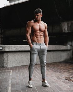 a shirtless man standing on a brick walkway with his hands in his pockets and looking at the camera