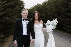 a bride and groom holding hands walking down a road in front of some pine trees