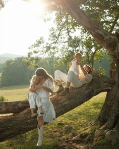 three women in white dresses sitting on a large tree branch with the sun shining behind them