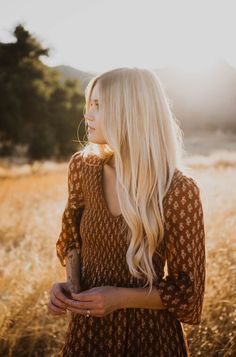 a woman with long blonde hair standing in a field looking off into the distance while wearing a brown and white patterned dress