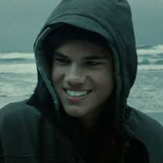 a young man with a hoodie on standing in front of the ocean and smiling