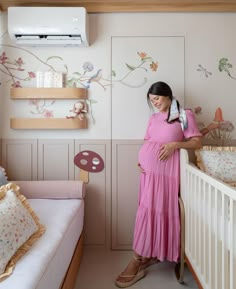 a pregnant woman in a pink dress stands next to her crib and looks at the camera