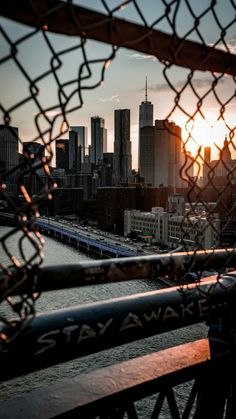 the sun is setting over a city skyline as seen through a chain link fence in front of a body of water