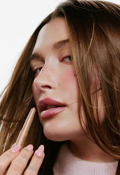 a woman with long brown hair and pink nail polish holding a lip balck in her right hand