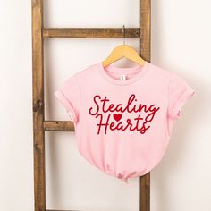 Looking for a cute tee for your kids? We have the perfect Stealing Hearts Cursive graphic tee addition to their closet! Also available in youth tees. Cute Cotton T-shirt With Heart Graphic, Cute Cotton T-shirt With Heart Shape, Cute Cotton T-shirt With Heart Design, Cute Heart-shaped Cotton T-shirt, Valentine's Day Graphic Cotton T-shirt, Pre-shrunk Cotton Top For Valentine's Day, Valentine's Day Cotton Tops With Letter Print, Cotton Pre-shrunk Tops For Valentine's Day, Valentine's Day Graphic Tee With Slogan