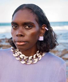 a woman with black hair wearing a purple shirt and statement earrings on her neck stands in front of the ocean