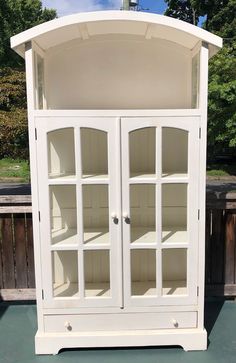 a white china cabinet with glass doors and shelves on the top shelf is sitting in front of a wooden fence