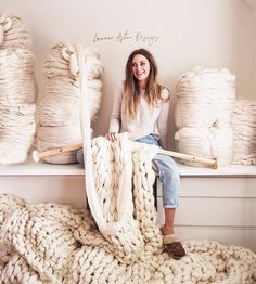 a woman is sitting on a bench holding a large knitted blanket in front of stacks of yarn