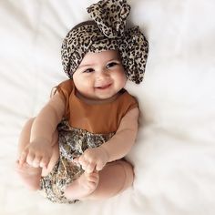 a baby wearing a leopard print headband smiles at the camera while sitting on a bed