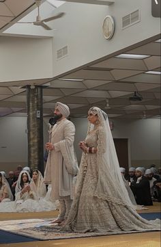 two people dressed in wedding attire standing on a stage with other people sitting down behind them