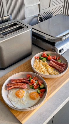 two plates with eggs, ham and vegetables on a cutting board next to an electric toaster