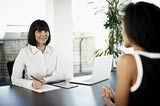 two women sitting at a table in an office talking to each other and one woman is smiling