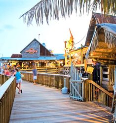 people are walking on the boardwalk near shops and restaurants at sunset or sunrise, with palm trees in the foreground