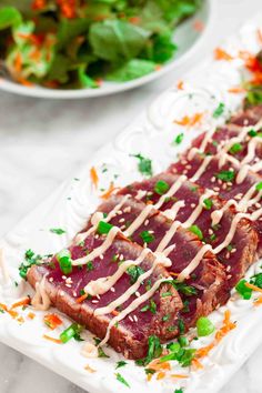 a white plate topped with meat covered in sauce and garnish next to a bowl of salad