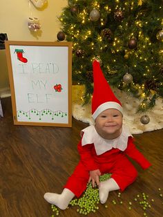 a baby dressed as elf sitting on the floor next to a christmas tree with peas