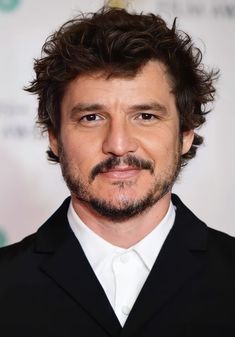 a man with curly hair and beard wearing a black suit, looking at the camera