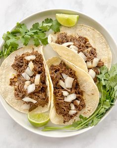 three shredded beef tacos on a plate with cilantro and lime wedges