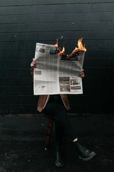 a person sitting on a chair holding up a newspaper with flames coming out of it