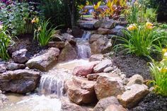 a small waterfall in the middle of a garden with lots of rocks and flowers around it