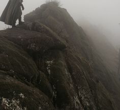 a person standing on top of a moss covered mountain in the foggy weather with a cape over their head