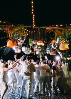 a large group of people dancing in front of some hanging lights and palm trees at night