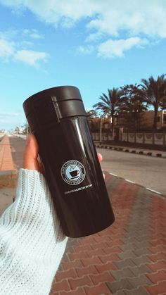 a person holding up a black coffee cup on the side of a brick road with palm trees in the background