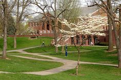 two people walking down a path in front of a brick building with white flowers on the trees