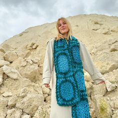 a woman wearing a blue crocheted scarf standing in front of rocks