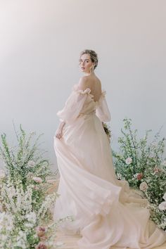 a woman standing in front of flowers and greenery wearing a long white dress with ruffled sleeves