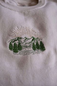 a white sweatshirt with green trees and mountains embroidered on it's chest, in front of the sun