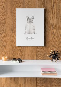 a white shelf topped with books and an art print on top of it next to a wooden wall