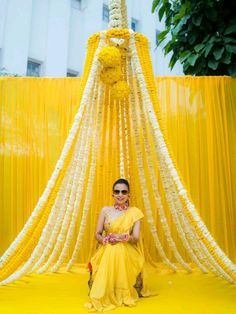 a woman sitting in front of a yellow and white structure with flowers hanging from it