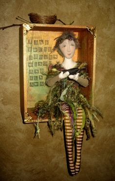 a doll is hanging on the wall in a frame with some plants and grass around it