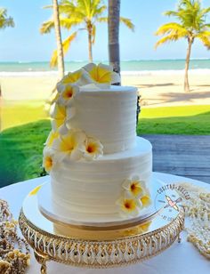 a white wedding cake with yellow flowers sitting on top of a table next to the ocean