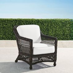 a wicker chair with white cushions sitting in front of a green hedge covered wall