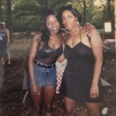 two women standing next to each other in front of trees and people on a field