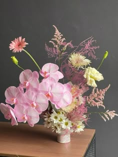 a vase filled with pink and white flowers on top of a wooden table next to a gray wall