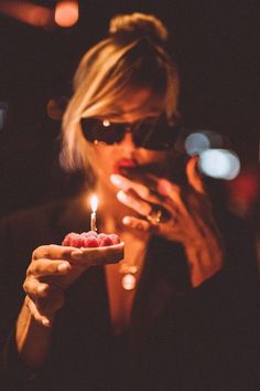 a woman holding a small piece of cake with a lit candle in her hand while looking at the camera