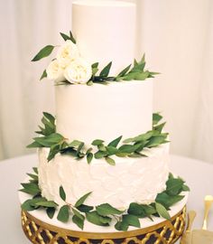 a white wedding cake with greenery on top and gold trim around the edges, sitting on a table